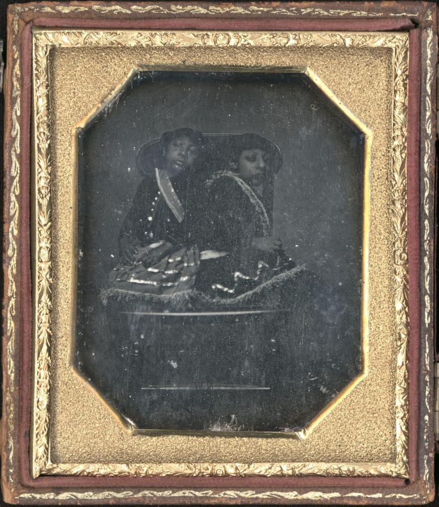 Daguerreotype of Maximo and Bartola, aka The Aztec Children. It is in a leather case with a gold frame and depicts the two children sitting on a chair together and dressed in their typical “Mesoamerican” style with a skirt and shirt combination. They are both very small. Details are difficult to make out because the background and lighting are both extremely dark.