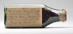 shriveled brown tea leaves sit inside a glass bottle that is closed up with a cork; cursive h和writing in brown ink on a yellowed paper is inside the bottle